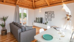A modern living room with a grey sofa, wooden ceiling beams, a TV on a white cabinet, and a dining table with glasses. - Indulge Porto Flats (Porto)