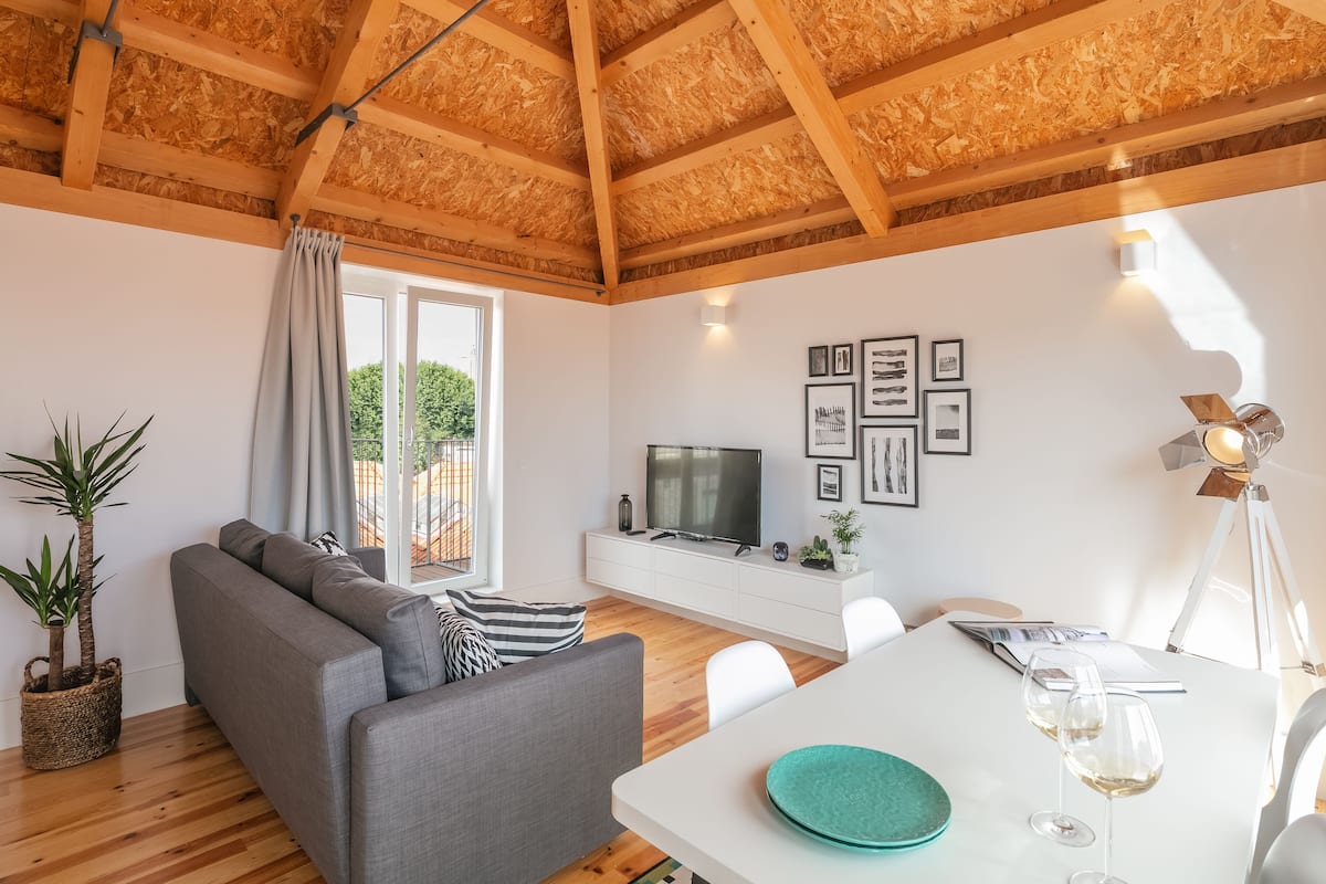 Een moderne woonkamer met een grijze bank, houten balken in het plafond, een televisie op een witte kast en een eettafel met glazen.