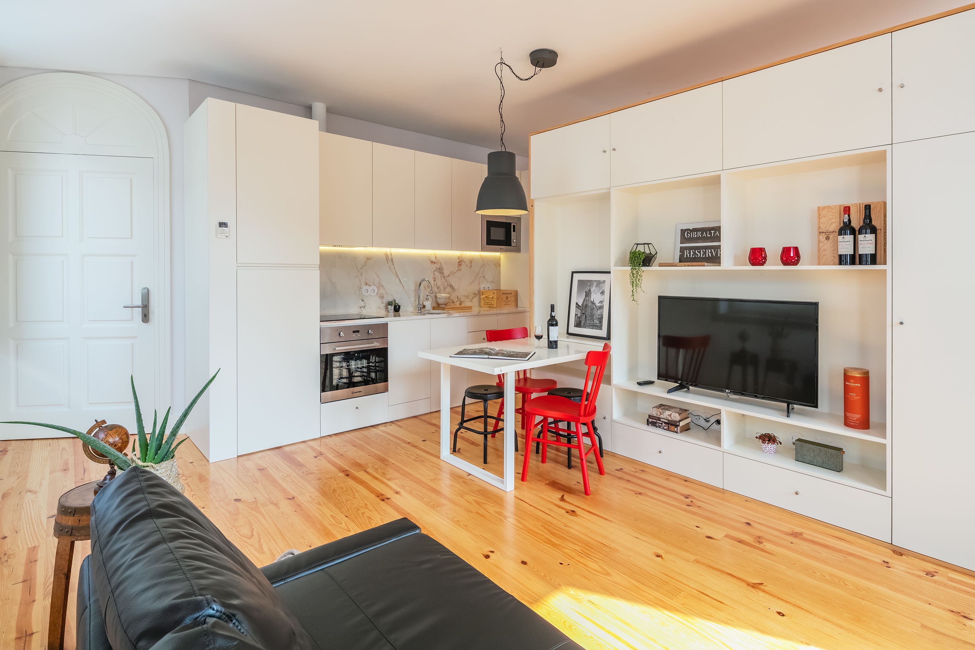 Una sala de estar moderna con un sofá negro, piso de madera, una zona de comedor con sillas rojas, una cocina con gabinetes blancos y un televisor sobre un estante blanco.