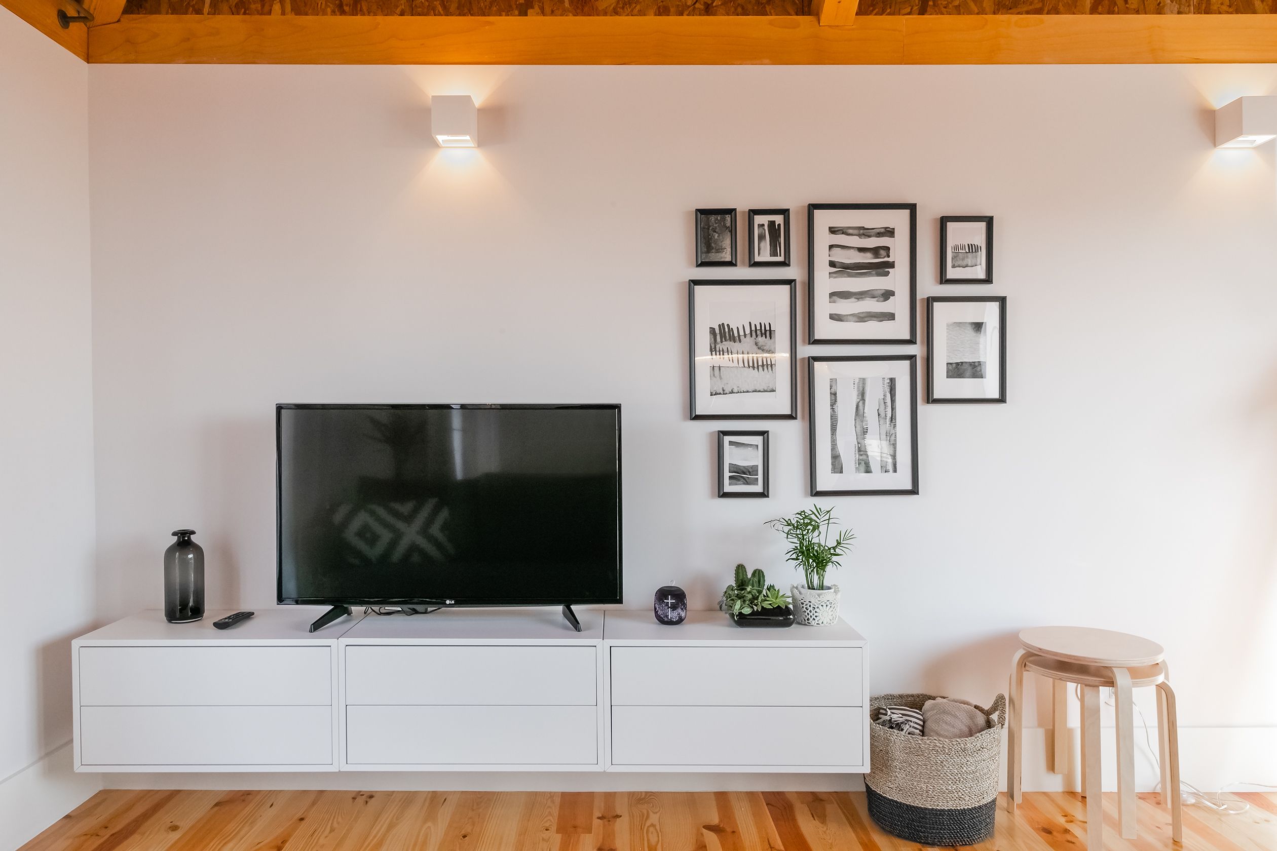 Ein modernes Wohnzimmer mit einem Flachbildfernseher auf einer weißen Kommode, einer kleinen Pflanze und gerahmten Kunstwerken an der Wand.