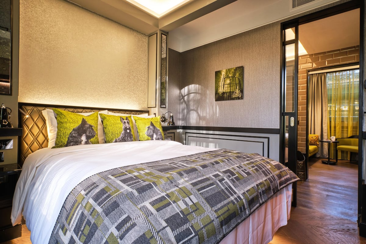 Une chambre d’hôtel avec un grand lit, une tête de lit ornée d’un motif géométrique, une œuvre d’art accrochée au mur et une vue sur l’extérieur par une porte.