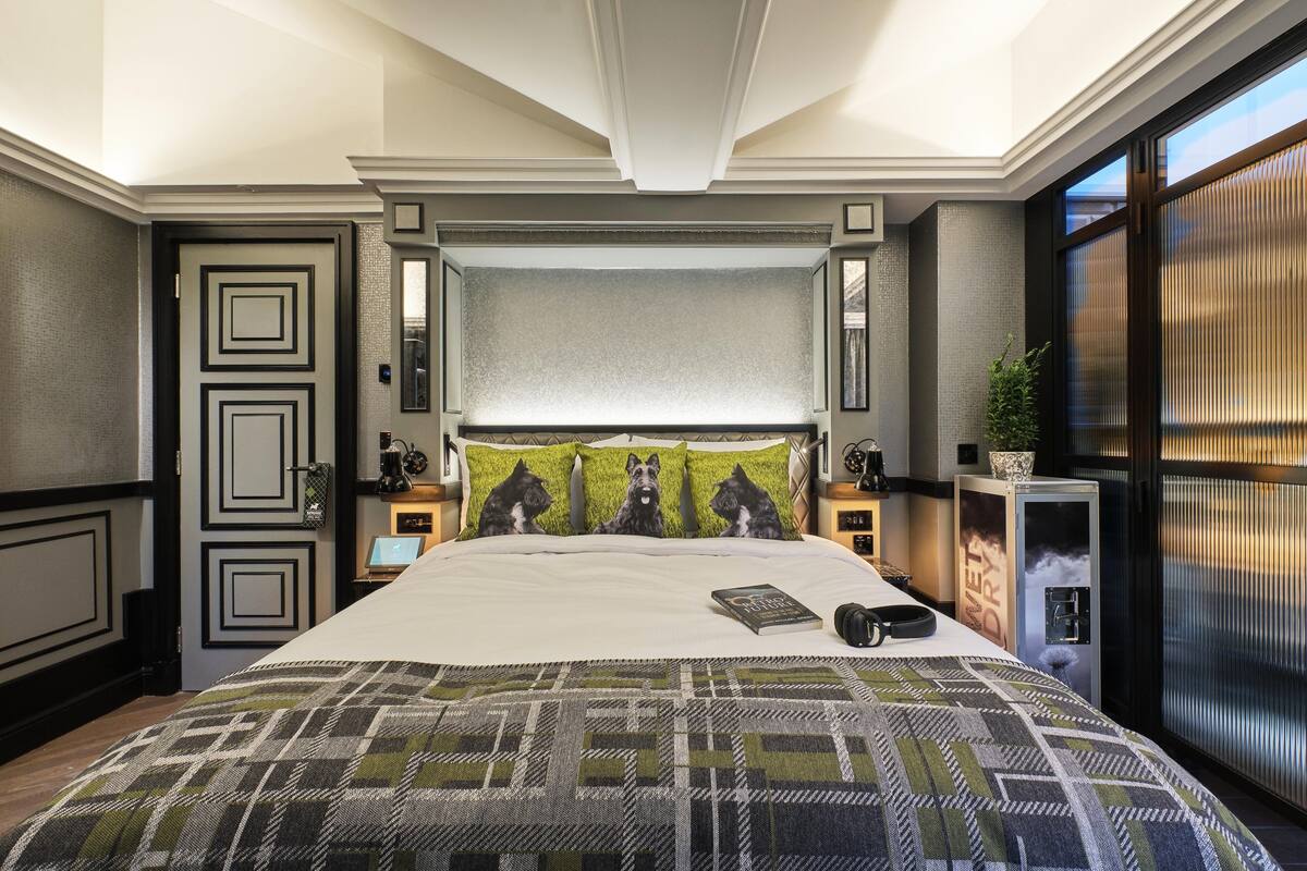 Une chambre à coucher avec un grand lit, une tête de lit agrémentée d’un miroir, une table de chevet avec une lampe et une fenêtre avec des stores.