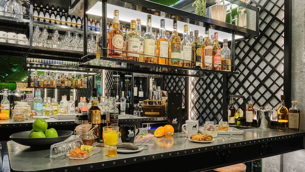Een bar met drankflessen op planken, een schaal groene appels, een glas sinaasappelsap en een bord croissants.