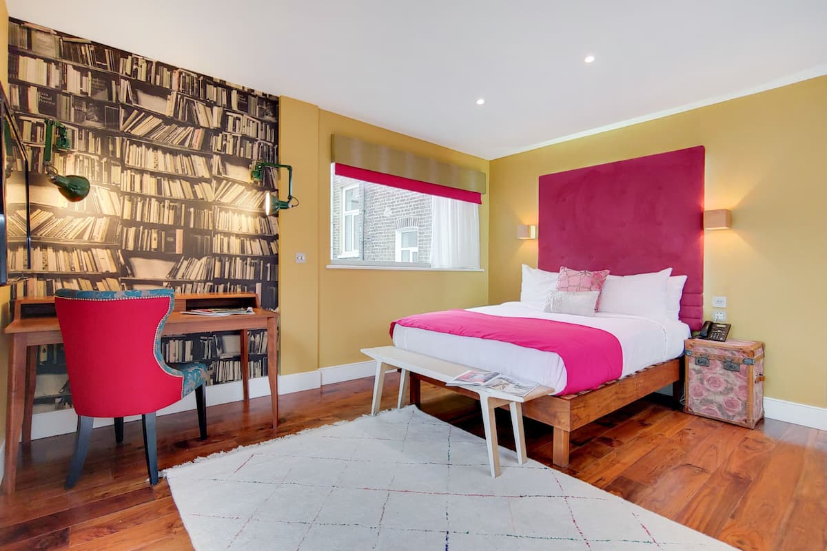 Une chambre avec un plancher en bois, une chaise rouge, un lit avec une tête de lit rose, une bibliothèque et une fenêtre avec un rideau rouge.