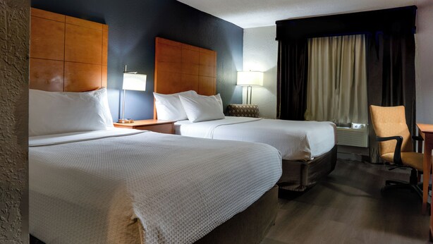 A hotel room with two beds, a wooden headboard, a nightstand, a lamp, a chair, and a window with curtains. - American Inn and Suites Ferndale (Ferndale)