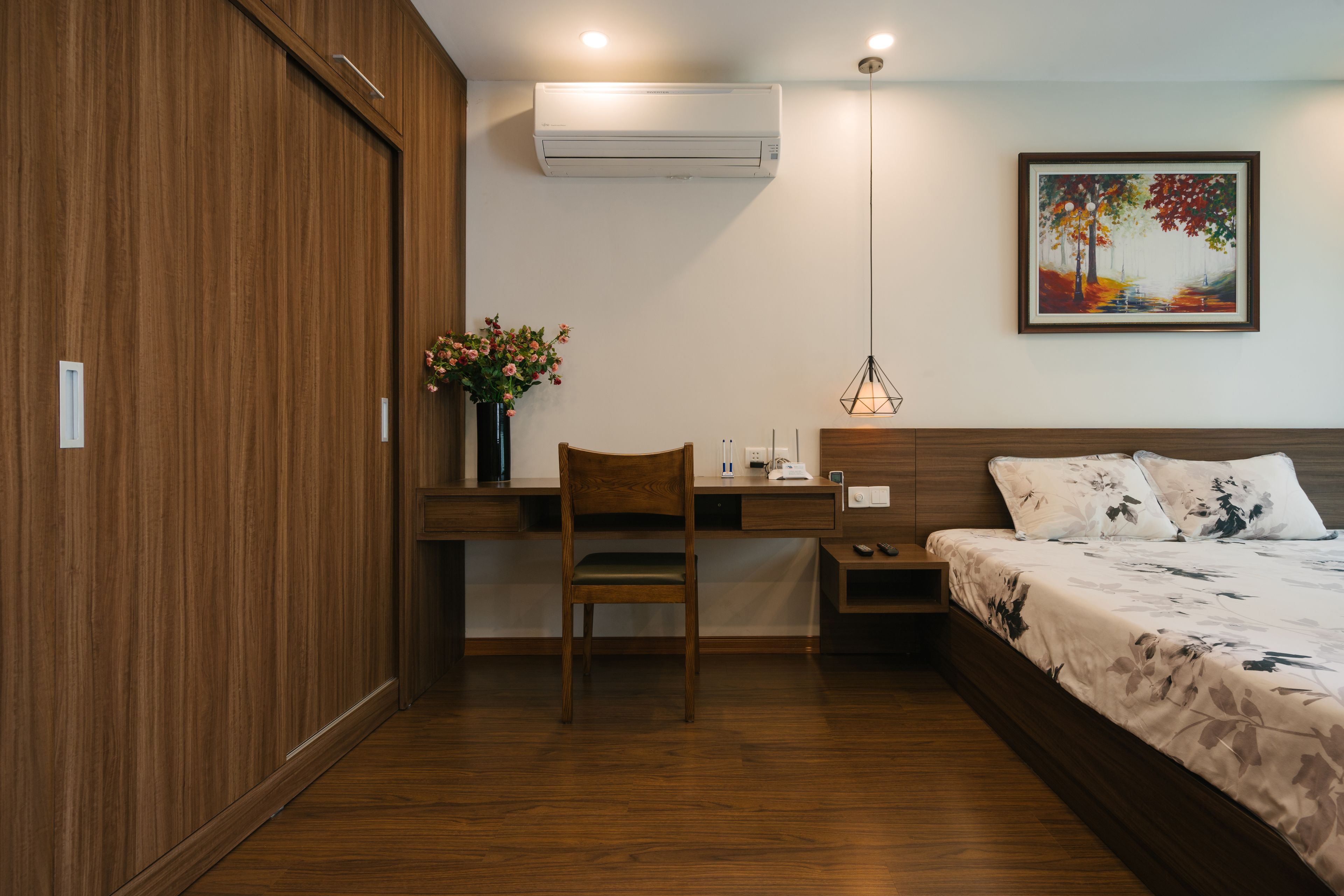 A bedroom with a wooden wardrobe, a desk with a chair, a bed with a floral bedspread, an air conditioner, and a painting on the wall.