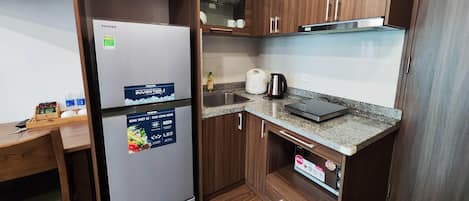 A compact kitchen with wooden cabinets, a stainless steel refrigerator, a countertop with a kettle, and a small sink.
