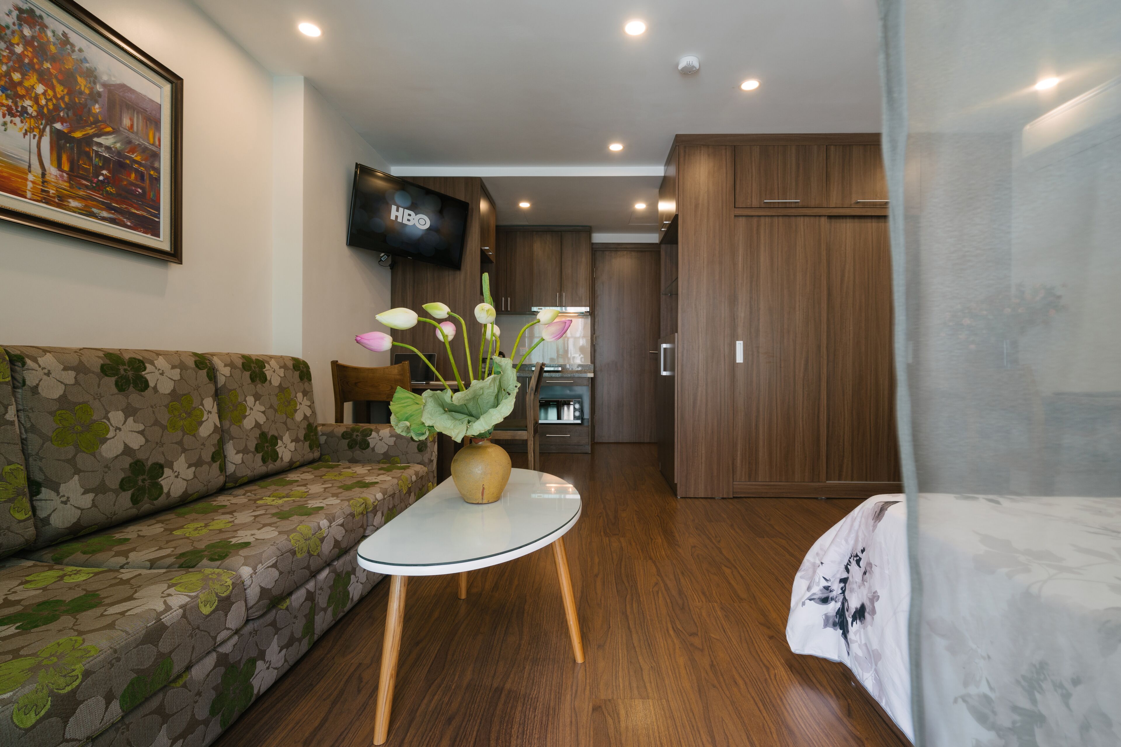 Una habitación de hotel moderna con sofá, una mesita redonda con un florero, piso de madera, televisor montado en la pared y un armario.