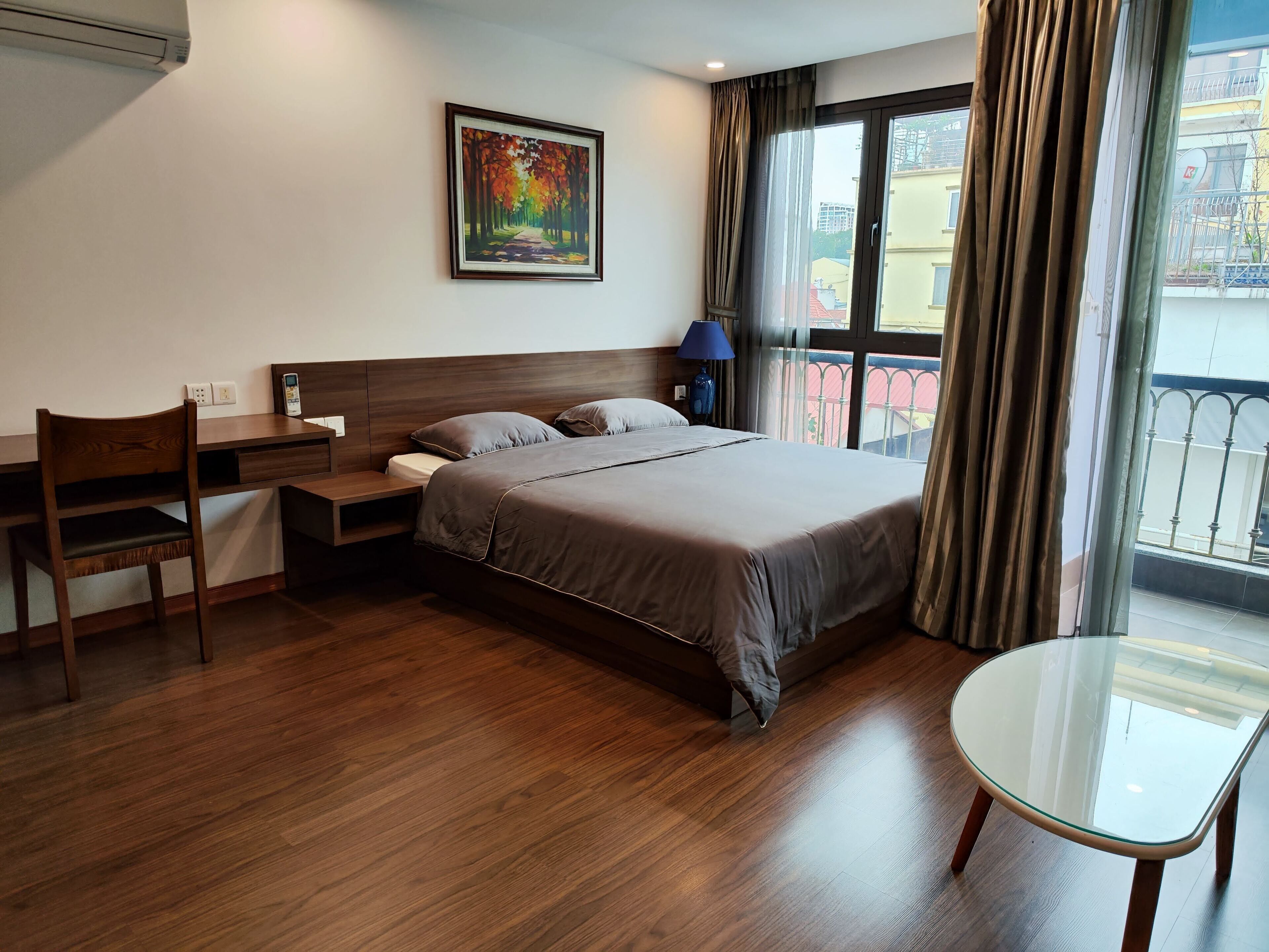 A hotel room with a bed, a desk, a chair, a glass table, a painting on the wall, and a view of the outside balcony.