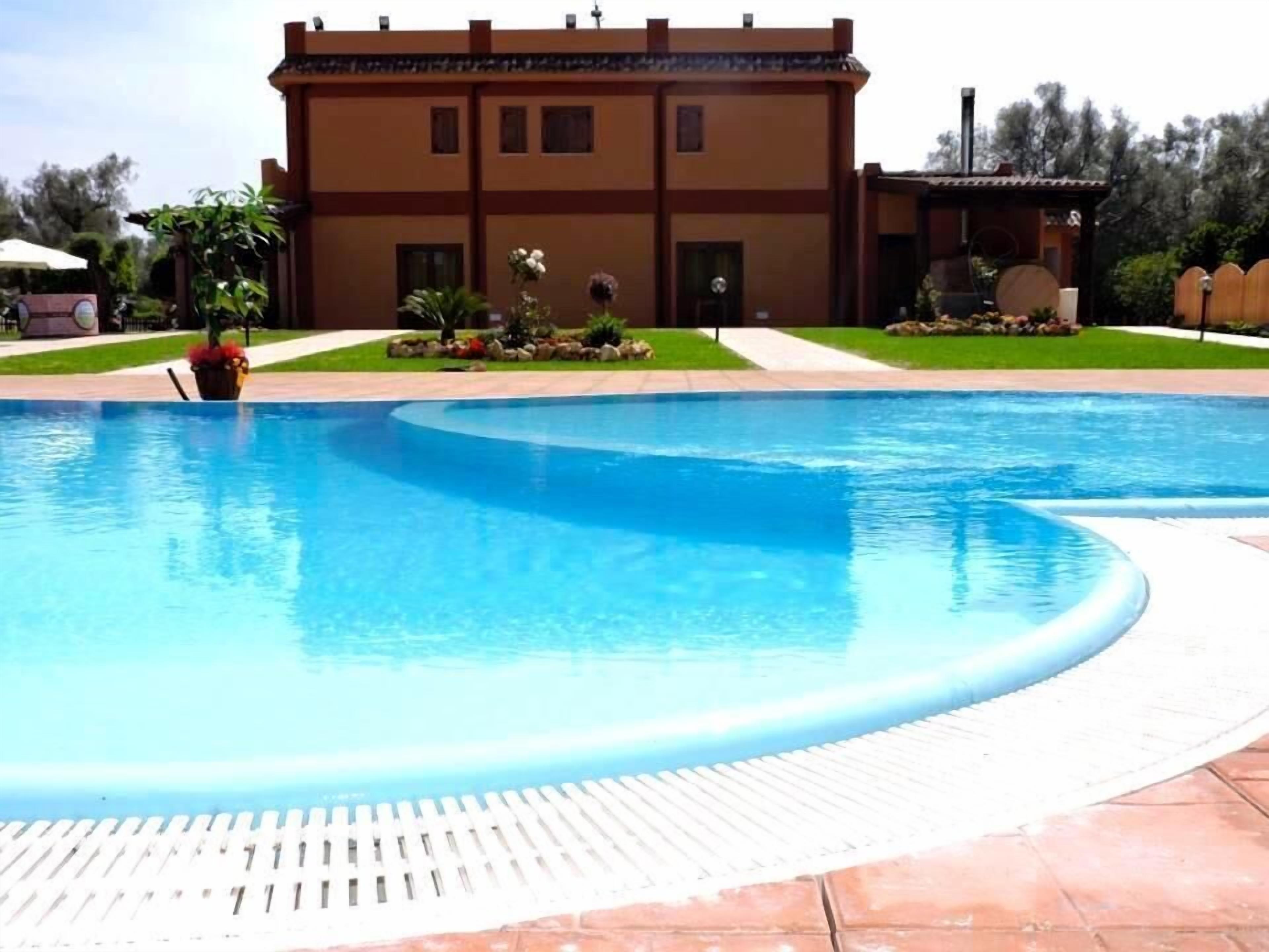 Une piscine à bord incurvé, entourée d’une terrasse carrelée. Au fond, on aperçoit un bâtiment à la façade marron et doté de nombreuses fenêtres.