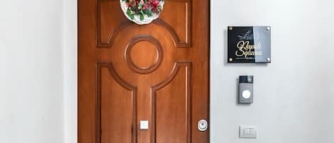 A wooden door with a wreath, a doorbell, and a mailbox. Below the door is a black mat with 'Napoli Squares SUITES' written on it.