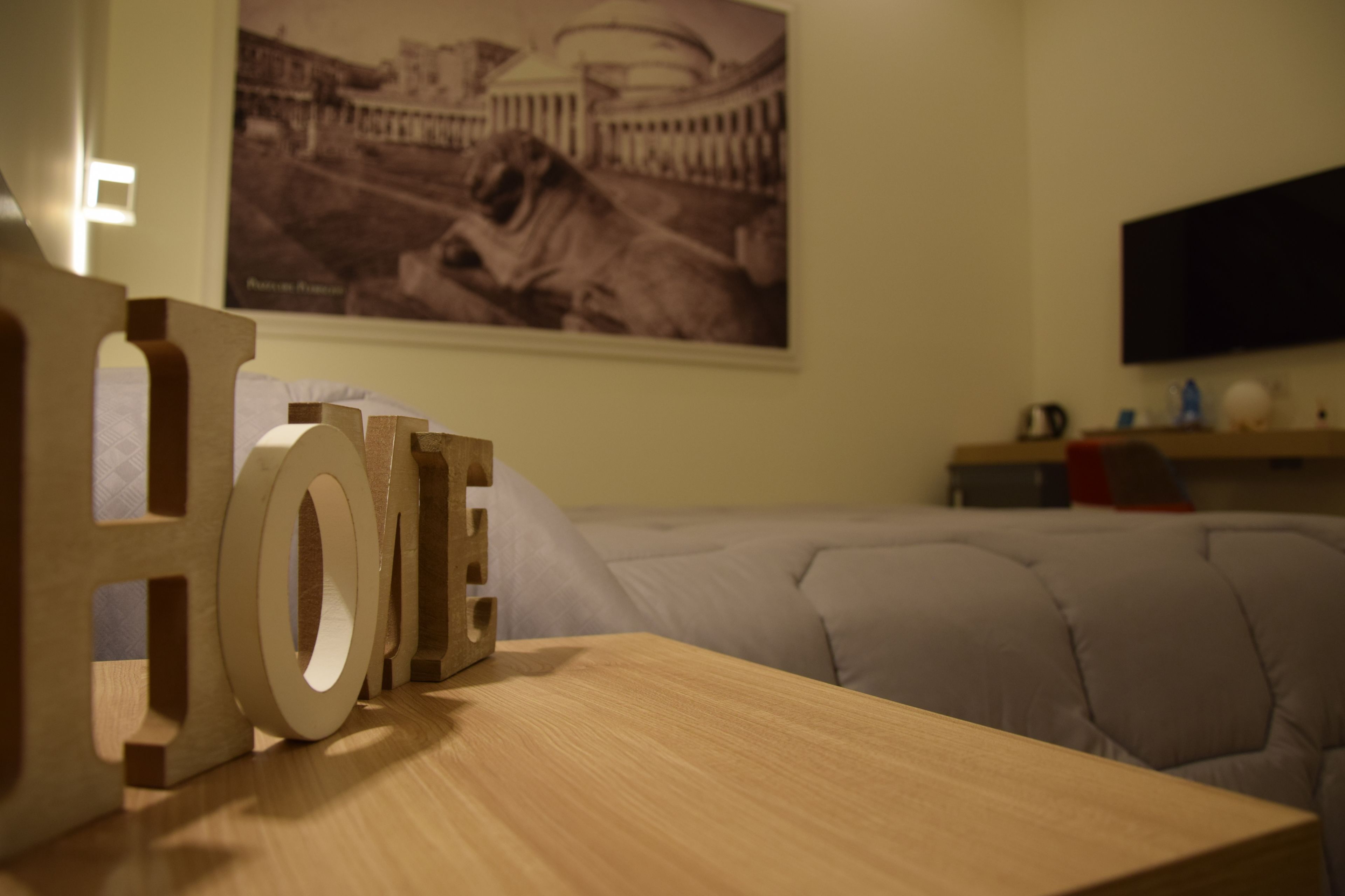 A room with a bed, a wooden table with letters spelling "HOME", a poster on the wall, and a television on a stand.