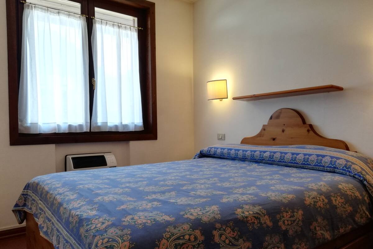 A bedroom with a bed, a window with curtains, a wall-mounted light, and a thermostat.