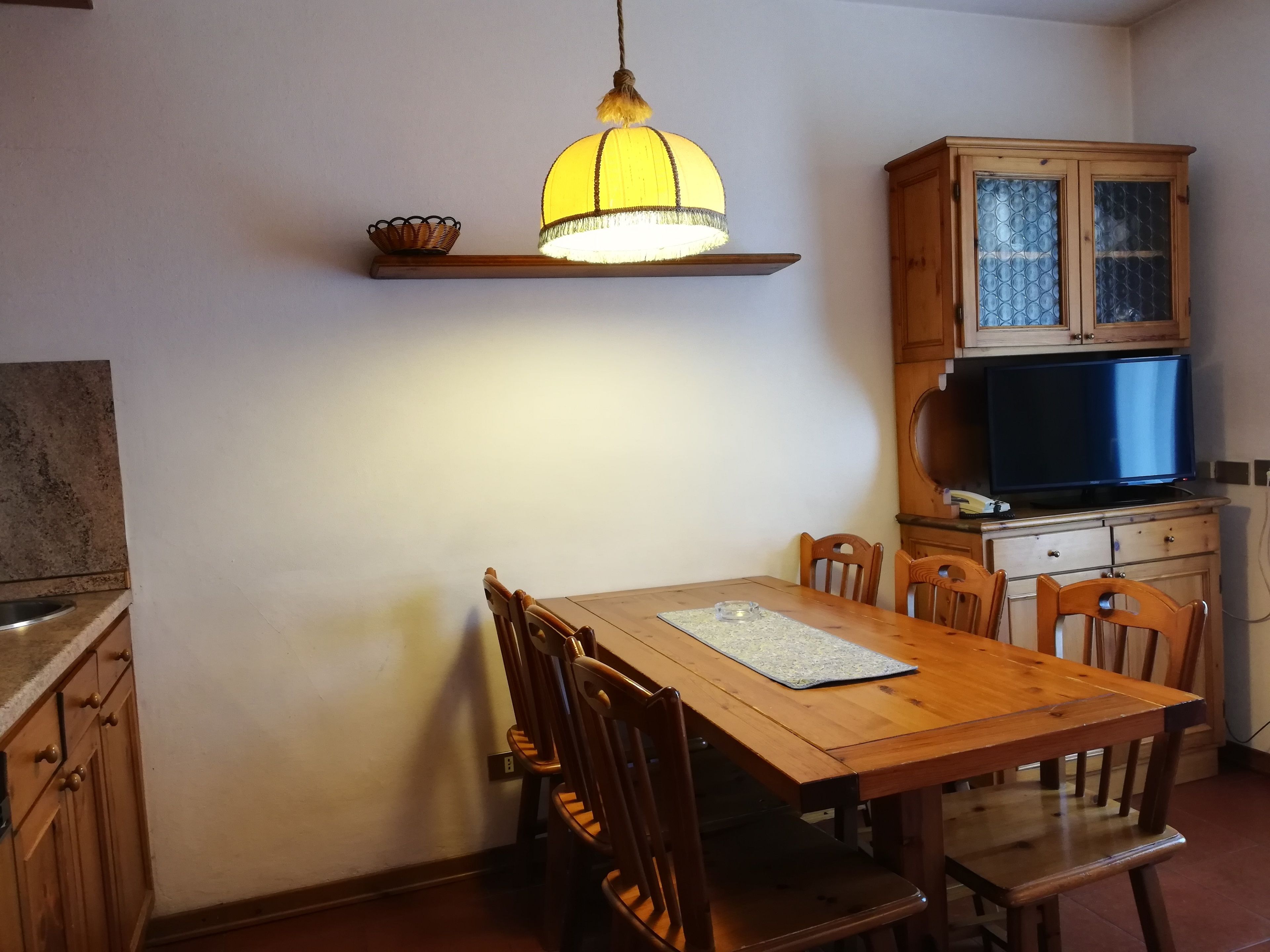 A dining area with a wooden table and chairs, a hanging lamp, a cabinet with glass doors, and a television on a wooden cabinet.