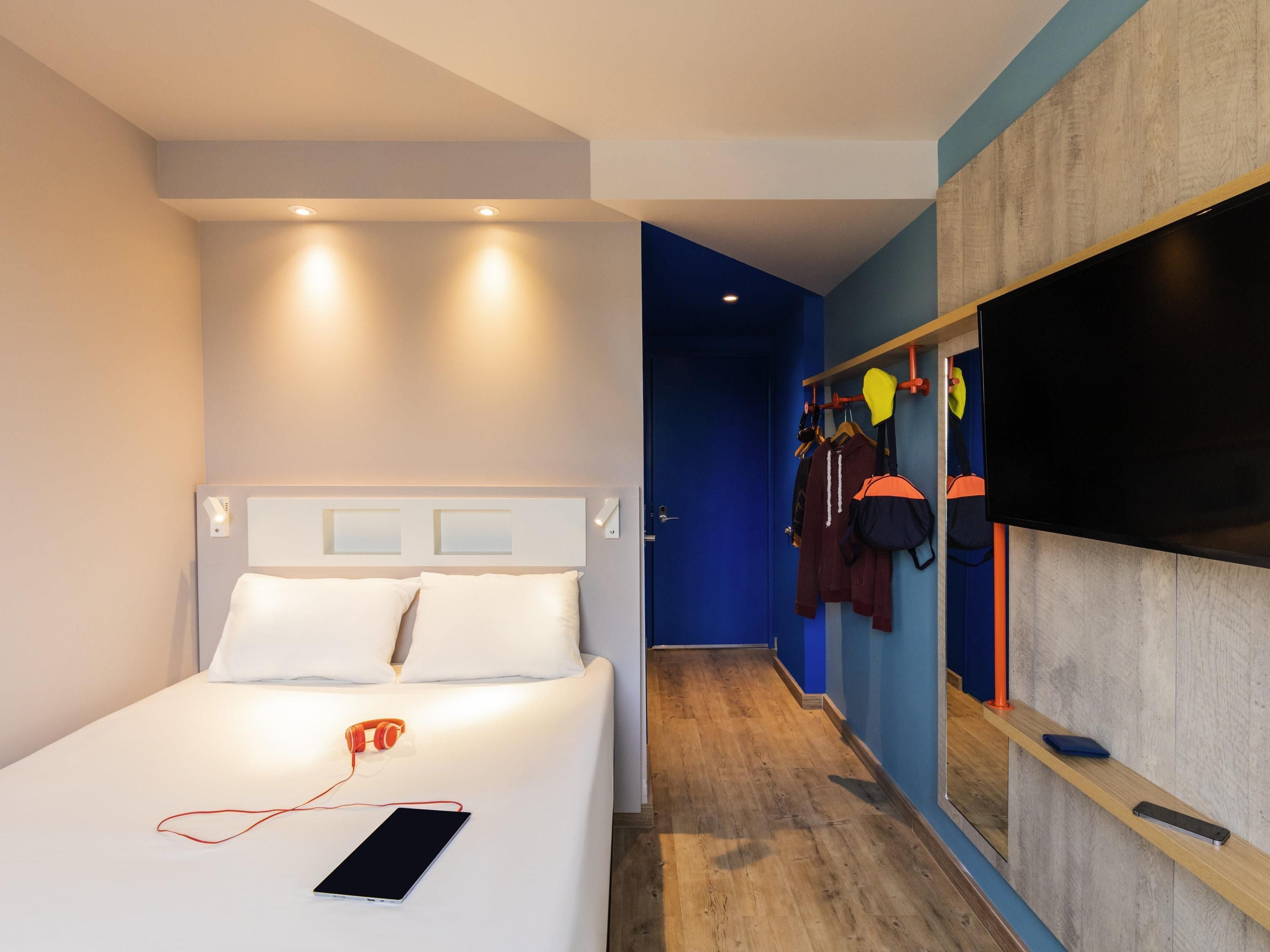 A modern hotel room with a bed, a bedside table with headphones and a tablet, a TV mounted on the wall, and a hallway leading to a closet with hanging clothes.
