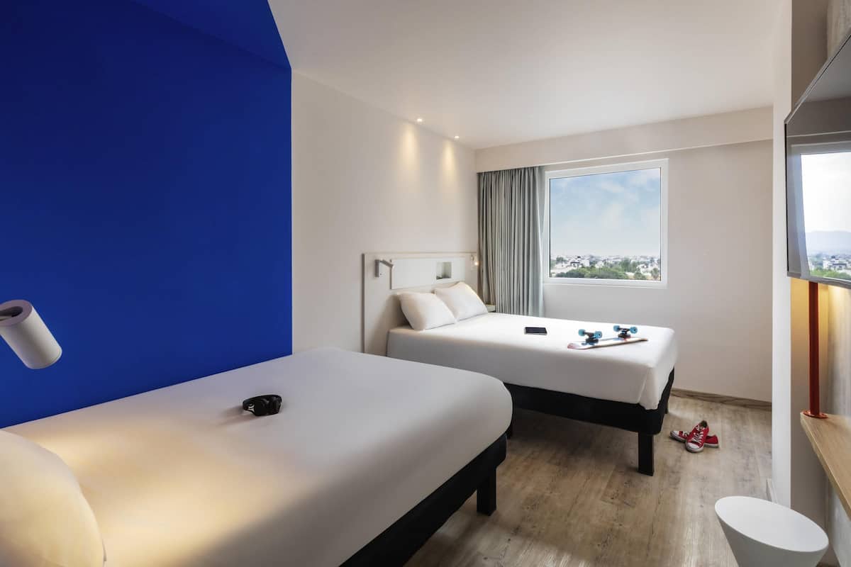 A hotel room with two beds, a large window with a city view, a desk with a chair, and a TV.