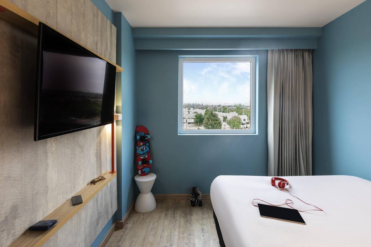 A hotel room with a TV mounted on the wall, a window with a city view, a bed with headphones, and a skateboard leaning against the wall.