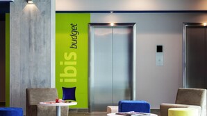 A modern lobby with a green accent wall, a central elevator, and comfortable seating areas with round tables and ottomans.