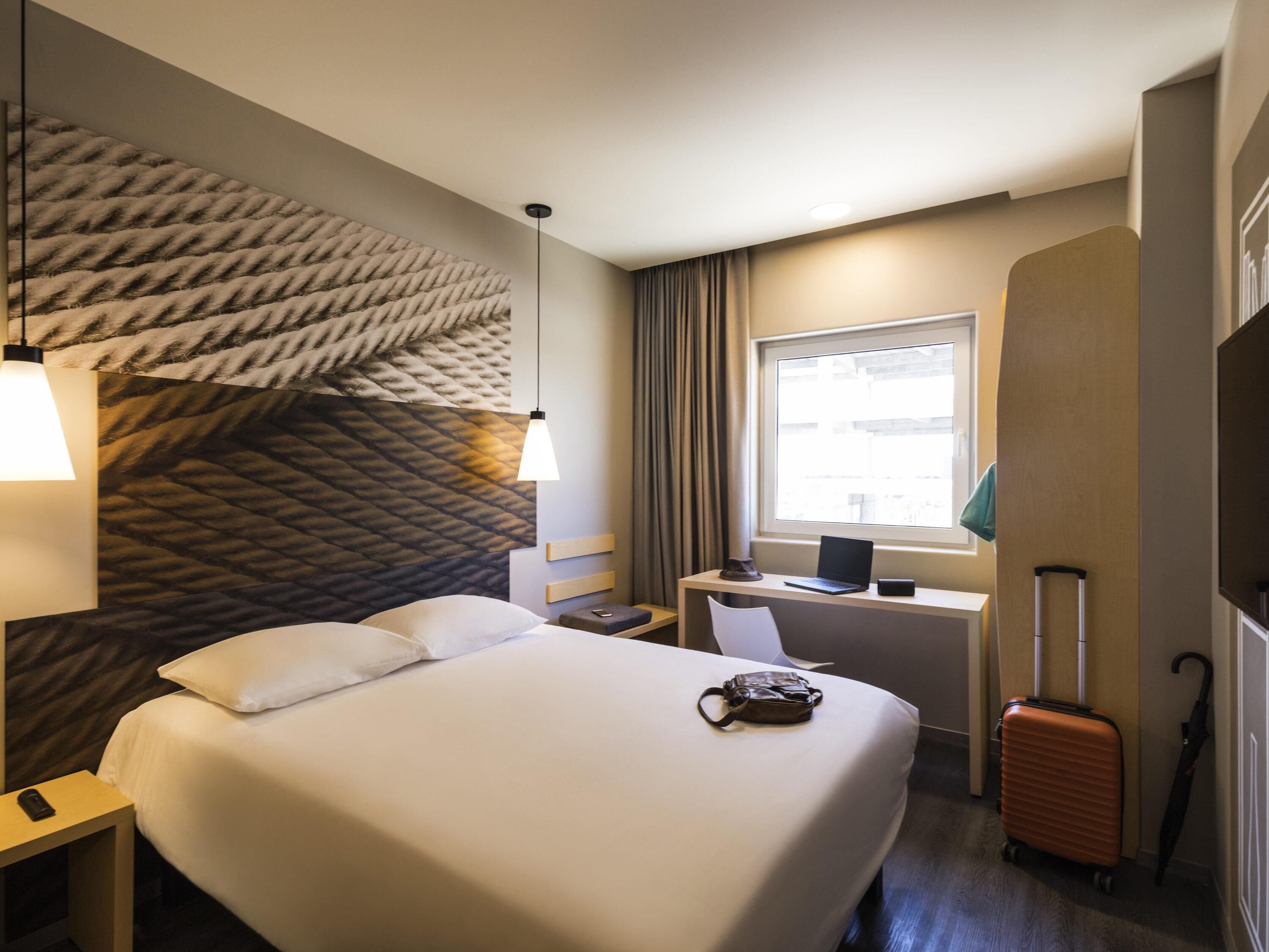 Una habitació d'hotel amb un llit gran, un escriptori amb un ordinador portàtil, una maleta i una finestra amb cortines.