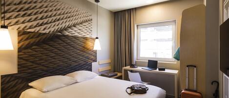 A hotel room with a large bed, a desk with a laptop, a suitcase, and a window with curtains.