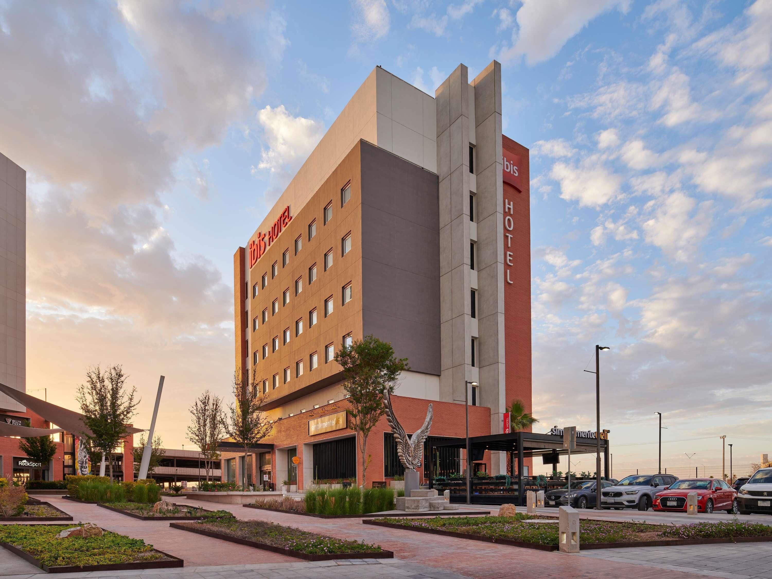 Un hôtel moderne, reconnaissable à son grand panneau « HÔTEL » sur la façade, doté d’un parking où sont stationnées plusieurs voitures et d’une cour aménagée avec des allées et de la verdure.