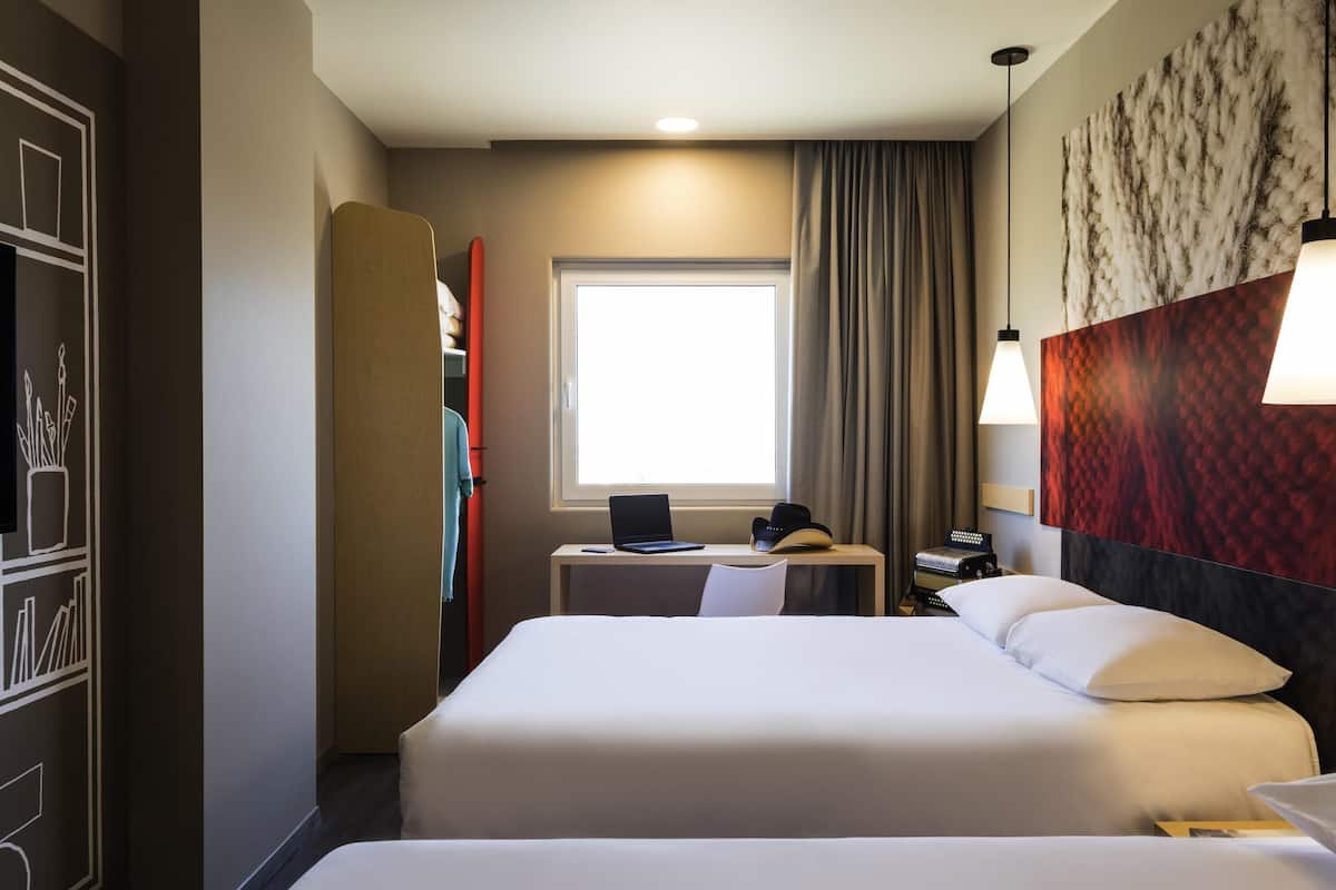 A hotel room with a bed, a desk with a laptop, a window with curtains, and a wall with a textured design.
