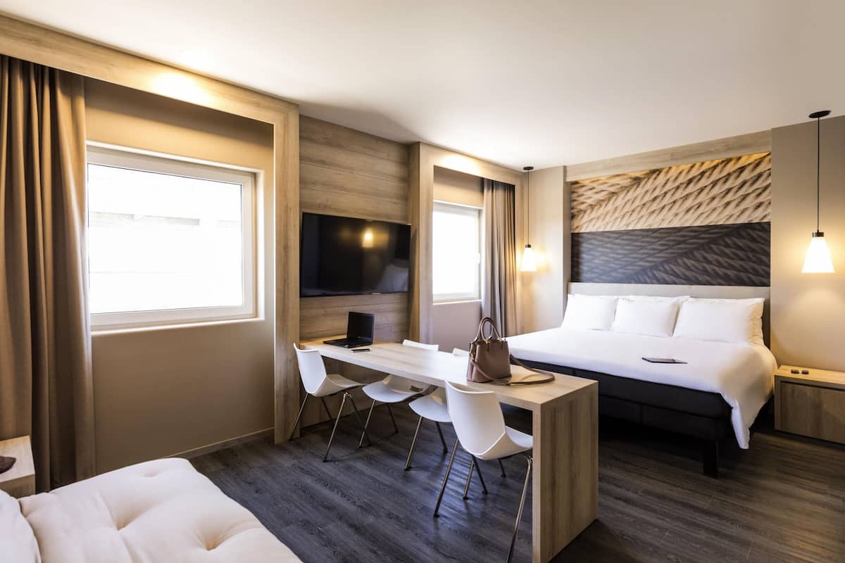 A modern hotel room with a bed, desk, chairs, and a TV.