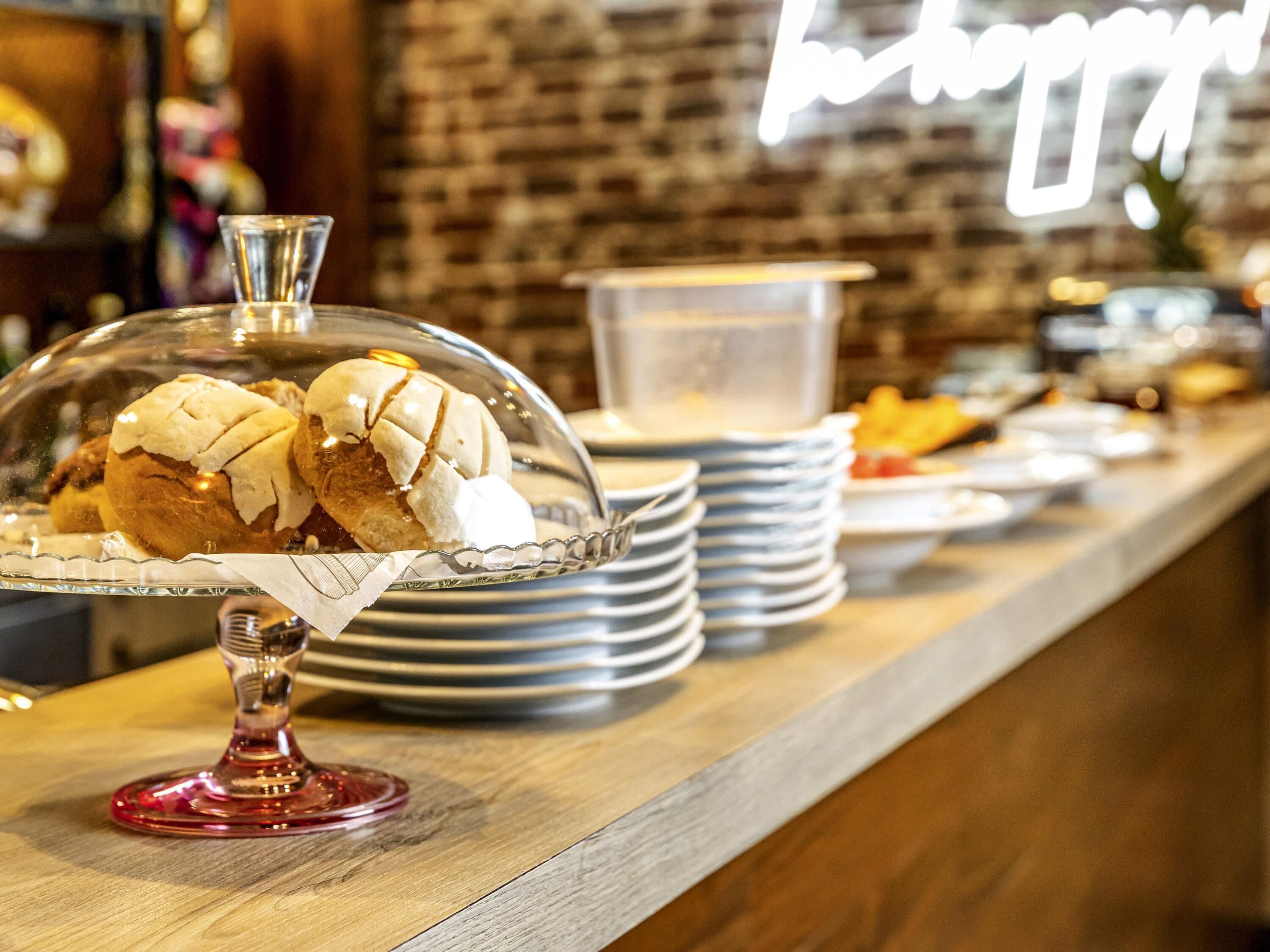 A breakfast buffet with pastries under a glass dome, stacked plates, and a neon sign in the background.