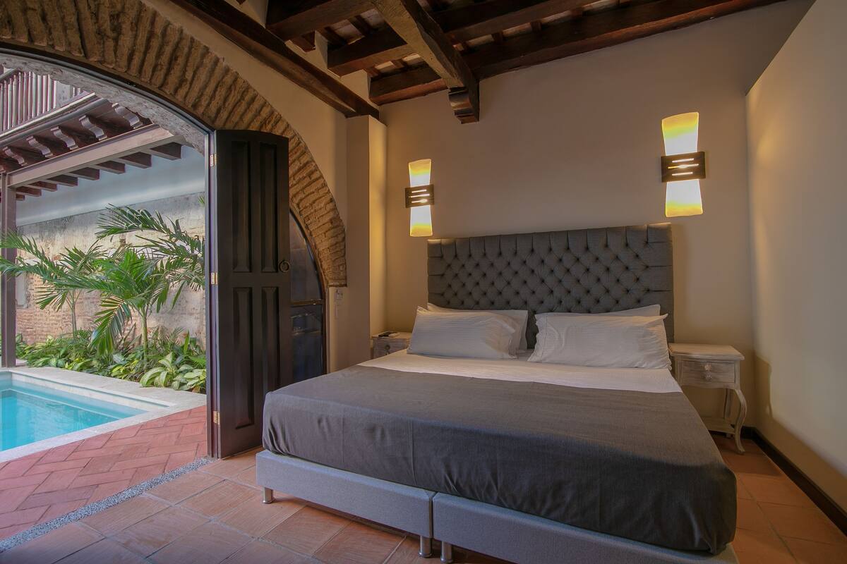 A bedroom with a large bed, a nightstand, a wall-mounted light, and a view of a swimming pool.