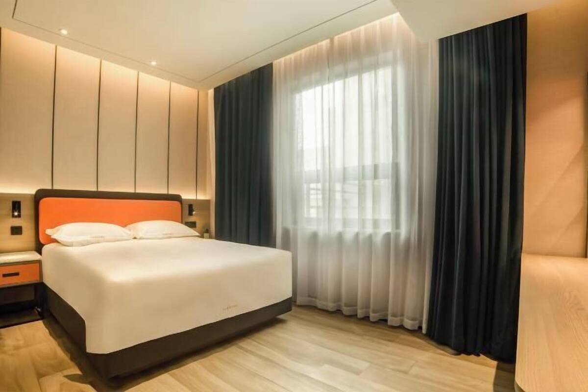 Ein modernes Hotelzimmer mit einem großen Bett, einem Nachttisch, einem Beistelltisch und einem Fenster mit durchsichtigen Vorhängen.