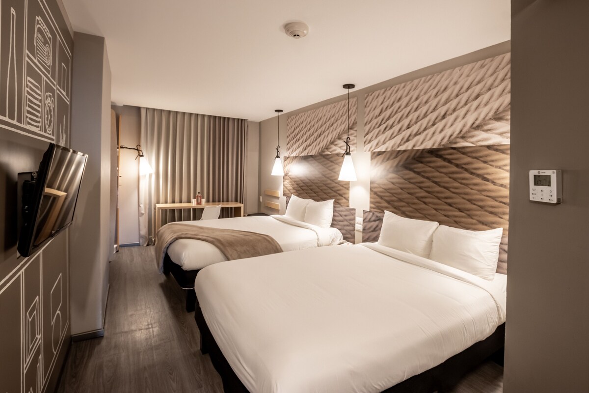 A hotel room with two beds, a TV, a desk, and a patterned headboard.
