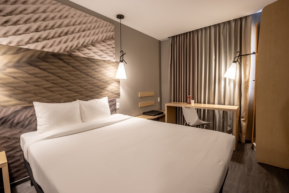 A hotel room with a large bed, a desk with a chair, a small table, and two pendant lights.