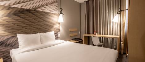 Una habitación de hotel con una cama grande, un escritorio con silla, una mesita y dos lámparas colgantes.