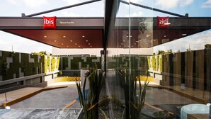 Une entrée d'hôtel moderne avec des portes en verre, une enseigne rouge et blanche indiquant « ibis HOTELS », et une vue dégagée sur la végétation et l'architecture environnantes.
