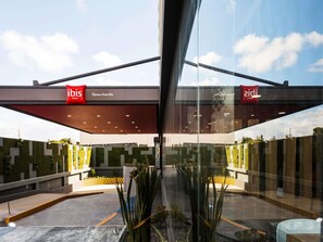 Una entrada de hotel moderna con puertas de vidrio, un letrero rojo y blanco que indica 'ibis HOTELS', y una vista despejada de la vegetación y la arquitectura circundantes.