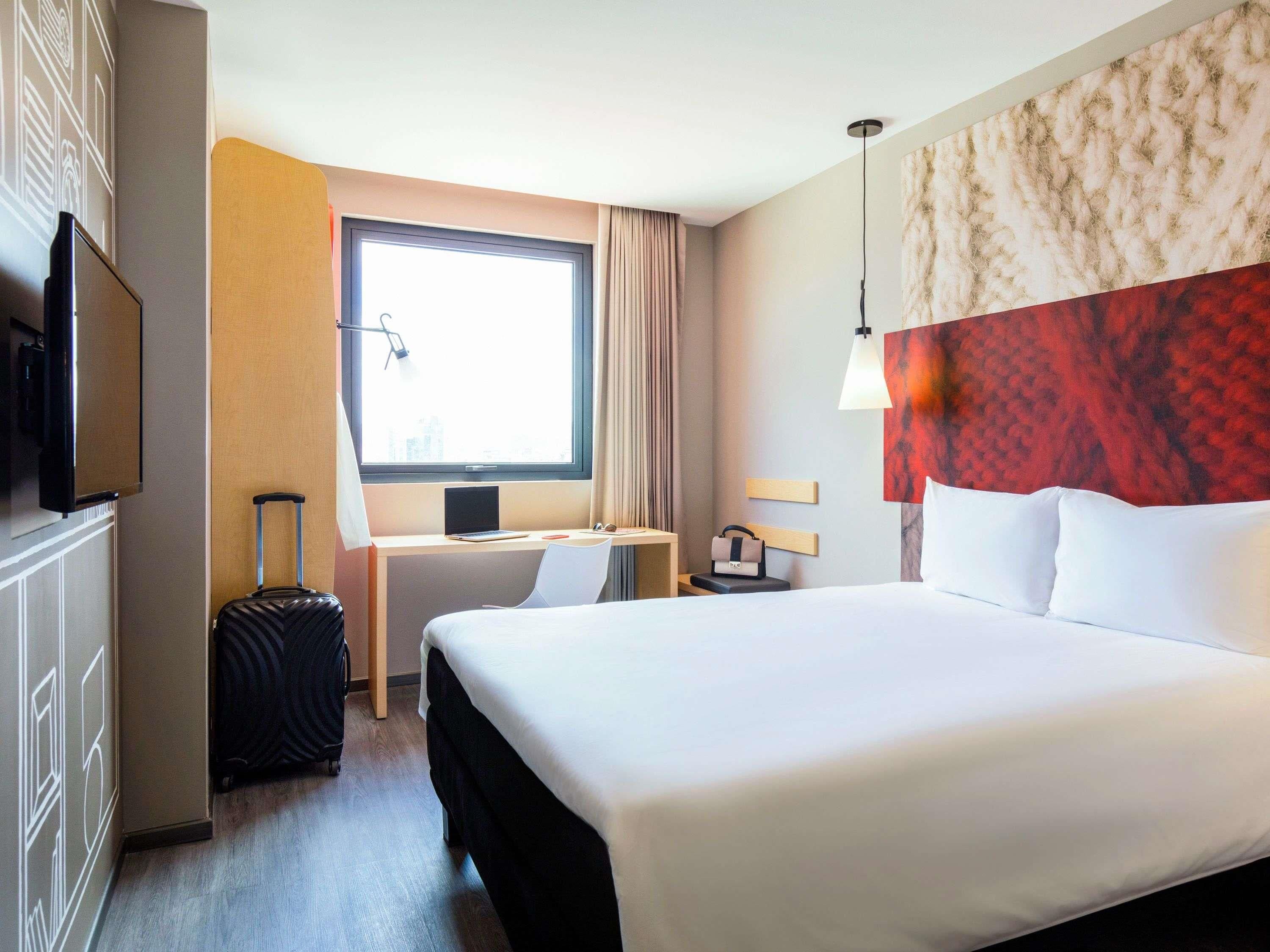 Habitación de hotel con cama, escritorio con computadora, televisor, maleta y ventana con cortinas.