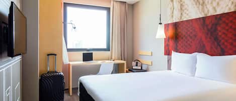 Habitación de hotel con cama, escritorio con computadora, televisor, maleta y ventana con cortinas.