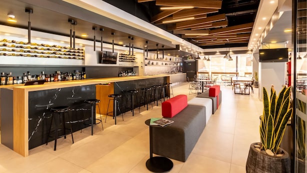 Un espace bar moderne doté d’un long comptoir, de plusieurs tabourets et d’un coin salon avec des coussins rouges.