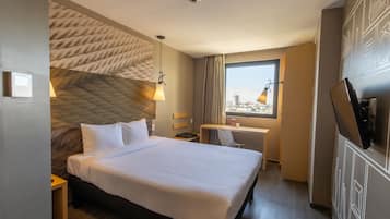 Habitación de hotel con una cama grande, un escritorio, una silla, un televisor y una ventana con vistas a la ciudad.