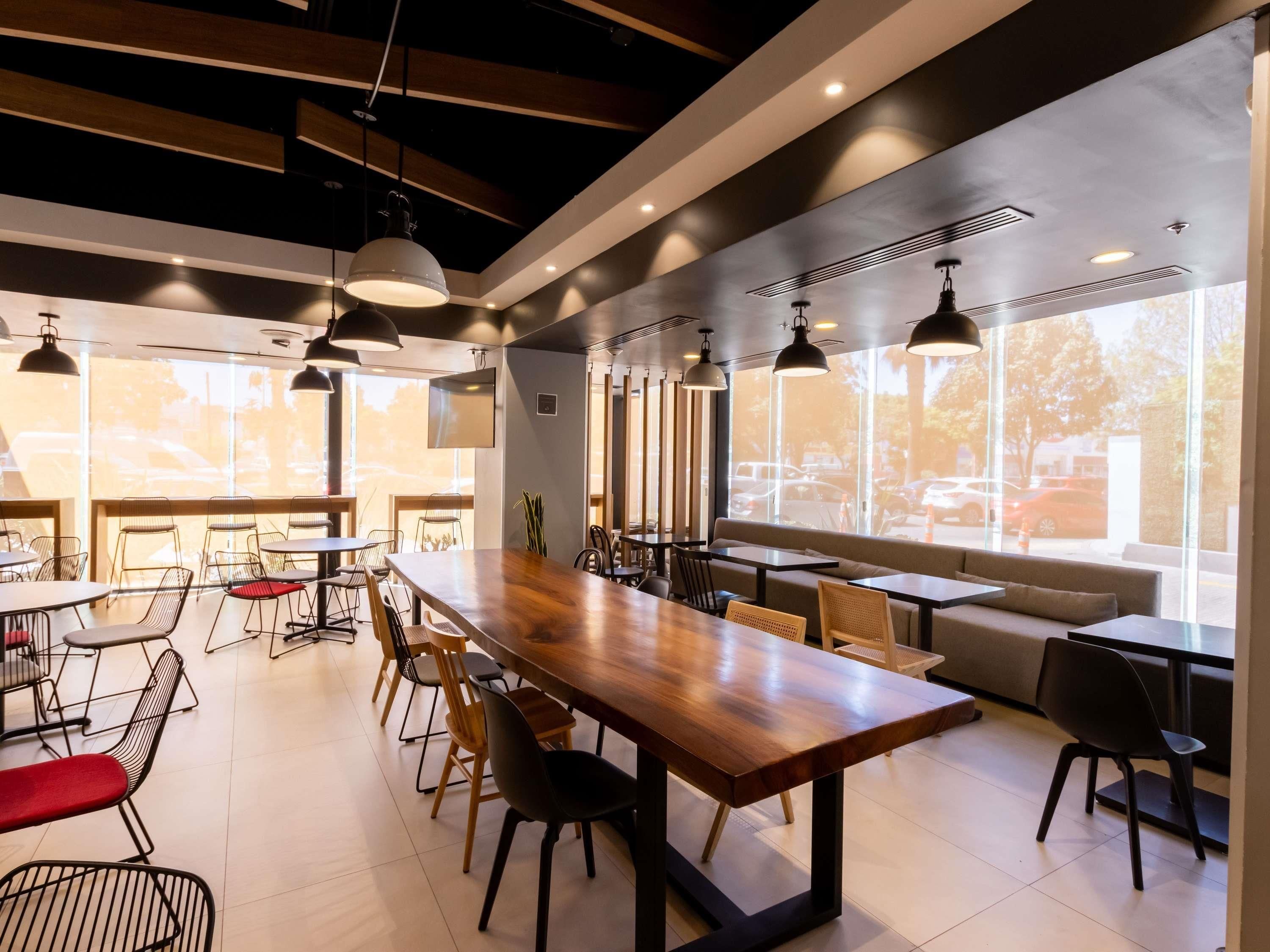 Un café moderno con una mesa de madera alargada, sillas negras e iluminación colgante.