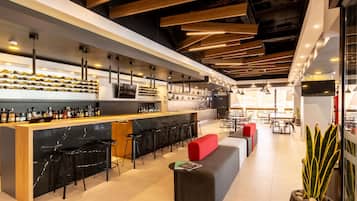 Un bar moderno con una barra larga, varios taburetes y un área de asientos con cojines rojos.