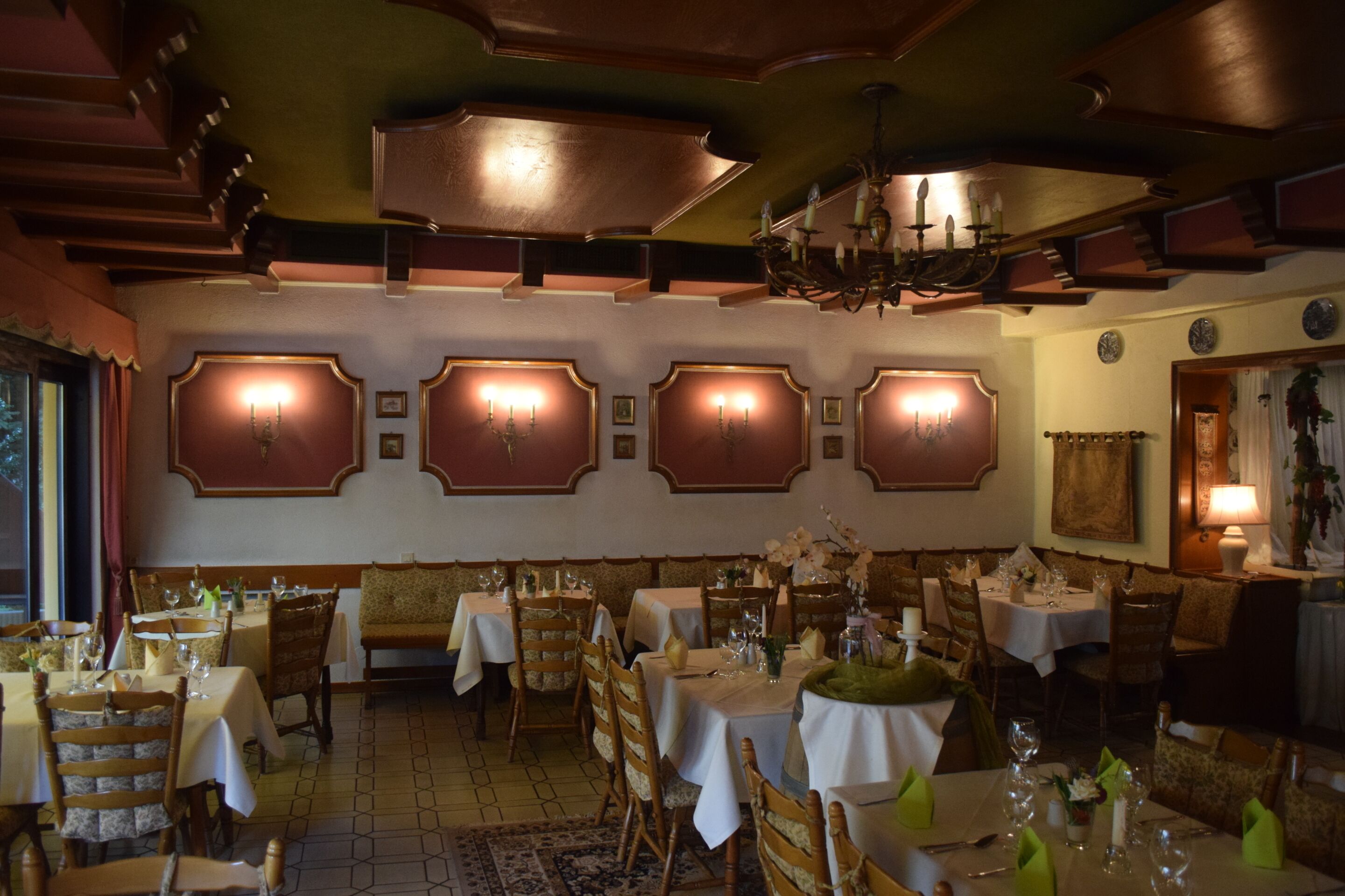 A restaurant with multiple tables and chairs, a chandelier, and wall-mounted sconces.