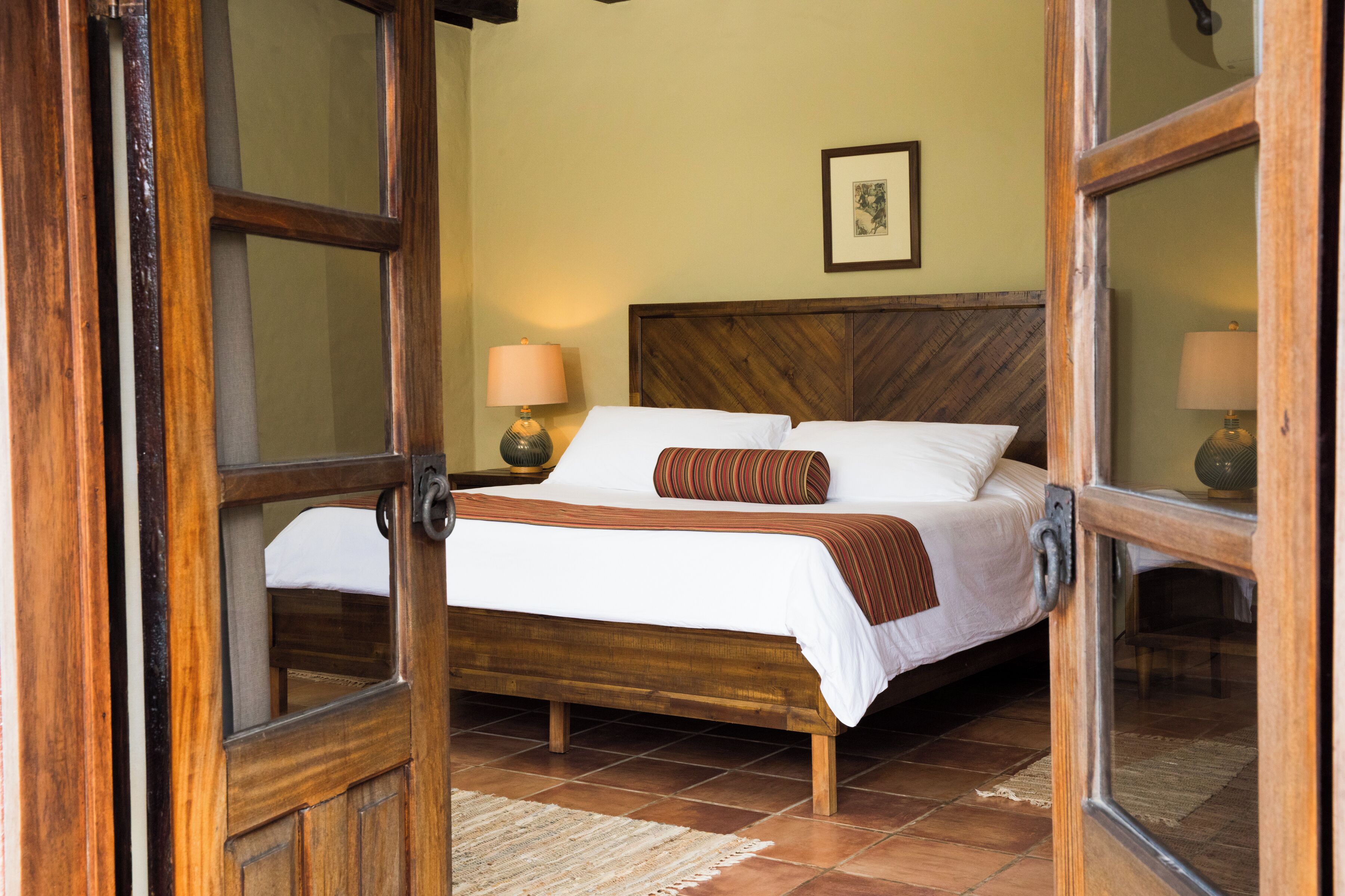 Una cama de madera con ropa de cama blanca y un plaid marrón, una mesita de noche con lámpara y un cuadro en la pared.