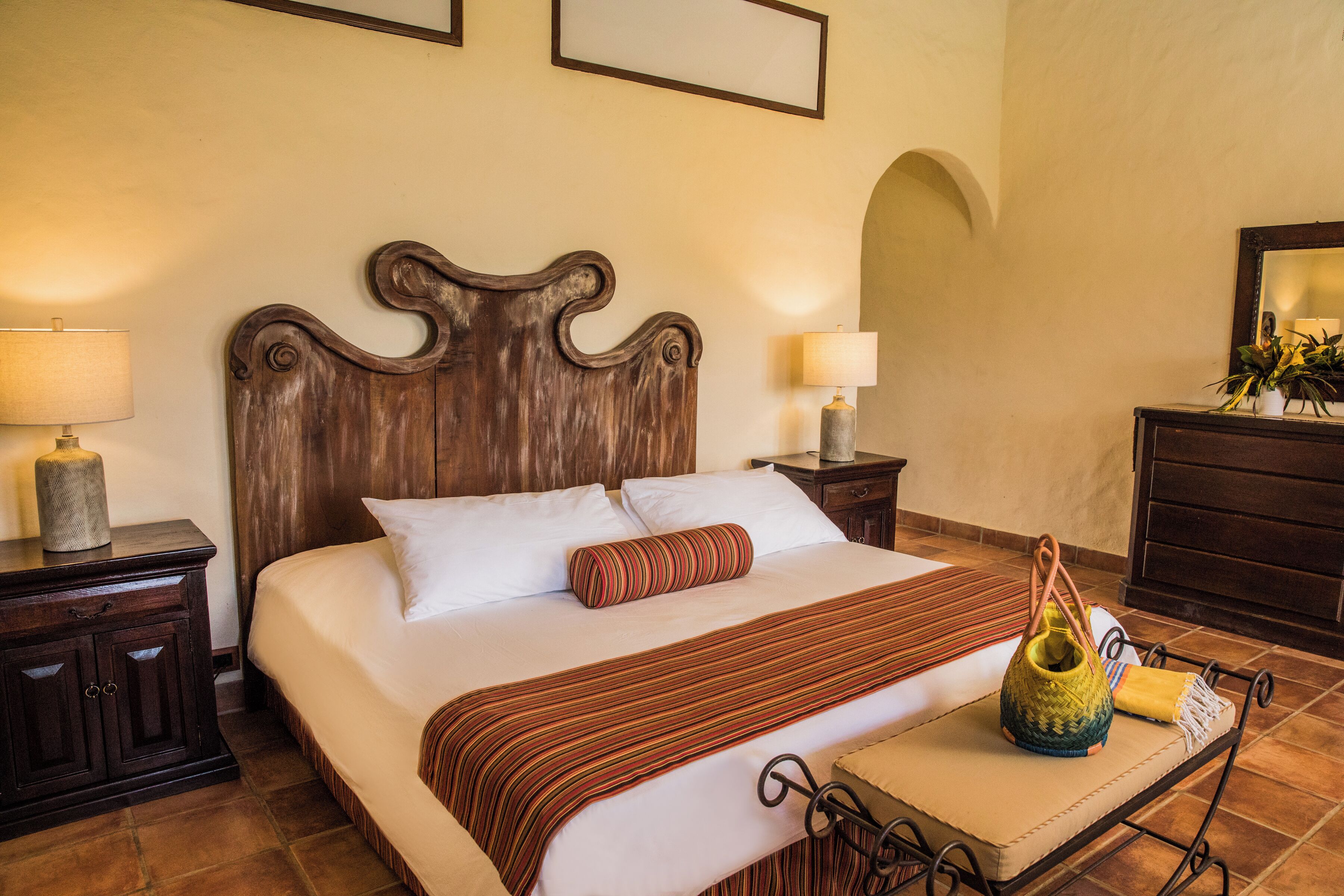 Una cama bien hecha con cabecera de madera, dos lámparas de noche, una mesita de noche con lámpara y una pequeña mesa con un adorno.