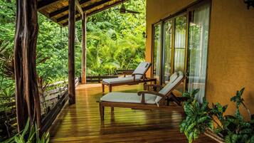 Una terraza de madera con dos sillones reclinables y una mesa, rodeada de exuberante vegetación y un ventanal grande.