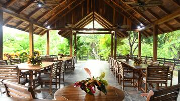 A covered outdoor dining area with wooden tables and chairs, surrounded by lush greenery.