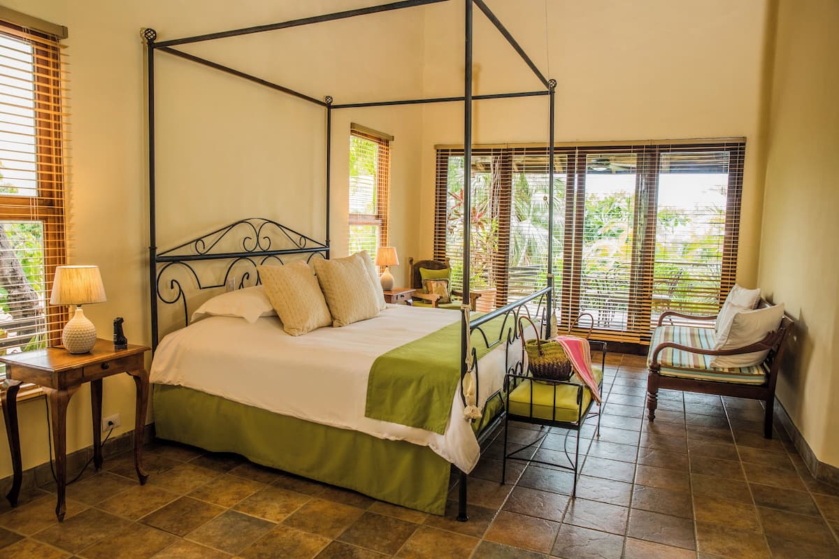 A four-poster bed with a green bedspread, a nightstand with a lamp, a chair, and a window with blinds.