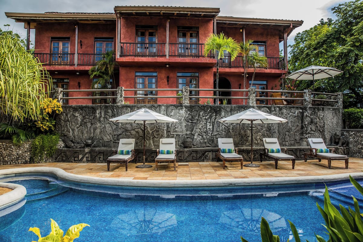 A pool area with lounge chairs and umbrellas, a stone wall with carvings, and a two-story building with balconies.