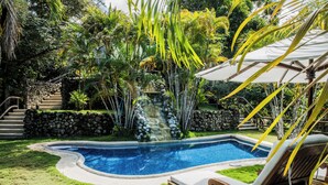 A swimming pool with a stone wall and a waterfall feature, surrounded by lush greenery and a lounge chair. - Hacienda Barrigona (Sámara)
