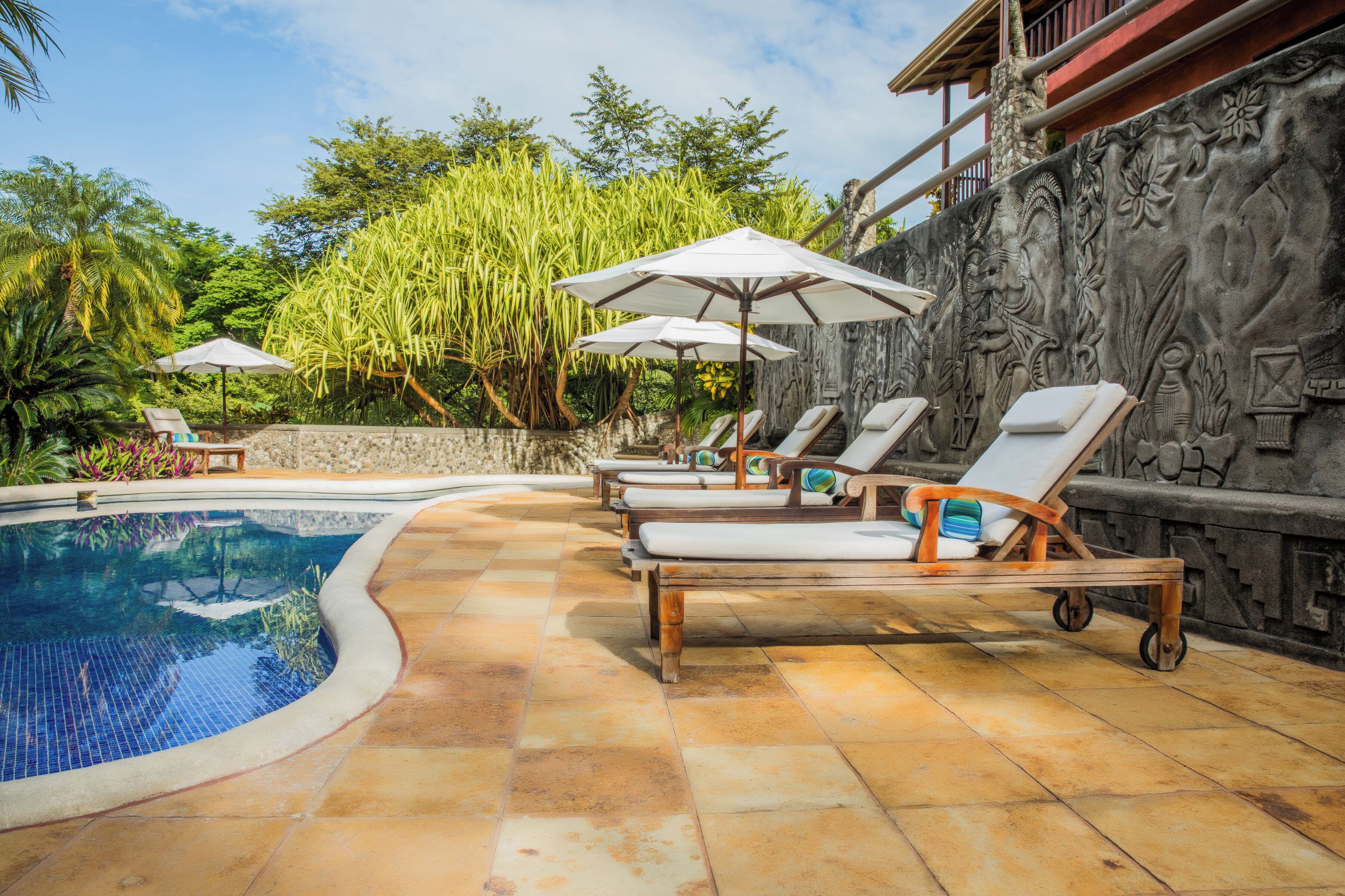 Un área junto a la piscina con sillones de descanso y sombrillas, rodeada de plantas tropicales y un muro de piedra decorativo.